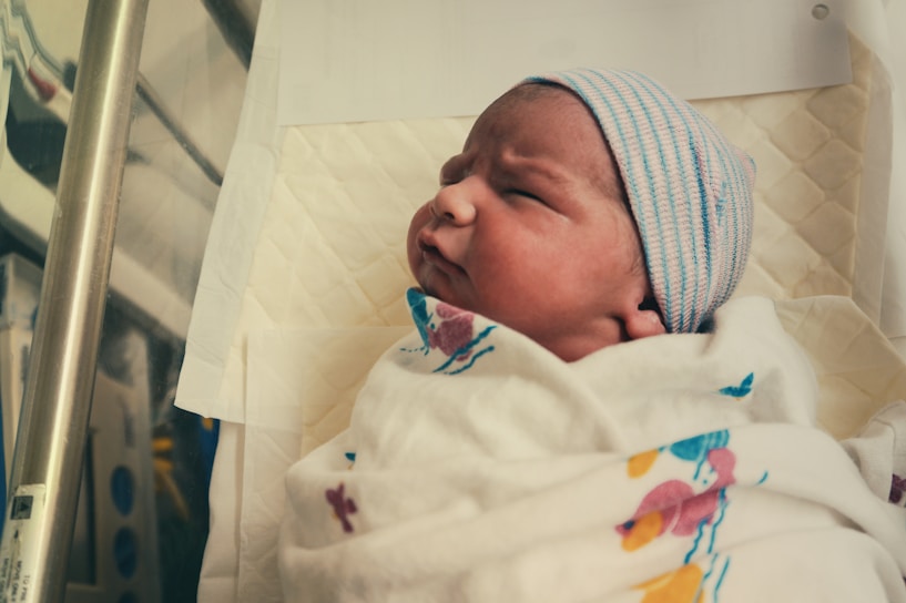 A newborn baby is wrapped in a white hospital blanket with colorful patterns. The baby is wearing a striped beanie and is lying on a padded surface.