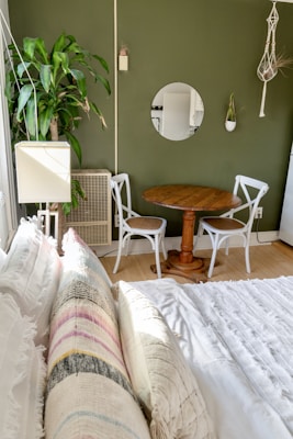 A cozy bedroom with a neatly made bed and layered pillows in pastel colors. To the side, there is a small wooden table accompanied by two white chairs. A large leafy plant is positioned next to a lamp and an air conditioner unit against an olive green wall. Above the table, a circular mirror and a small hanging plant in a macrame holder add decorative elements.