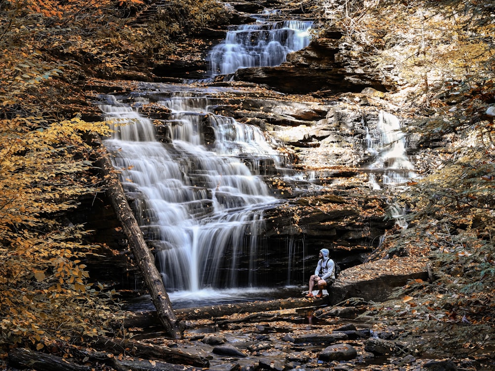 Hombre con chaqueta azul y pantalones negros de pie en la roca marrón cerca de las cascadas durante el día