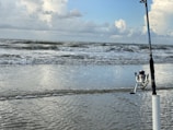 A fishing rod is standing upright on a sandy beach near the shoreline. The ocean waves are gently rolling in, and the sky is partly cloudy with various shades of blue and white.