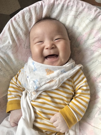 A smiling baby is lying on a blanket with a white and light pink pattern. The baby is wearing a white and yellow striped onesie with matching yellow cuffs. A light-colored cloth with blue and brown designs is draped over the baby's chest.