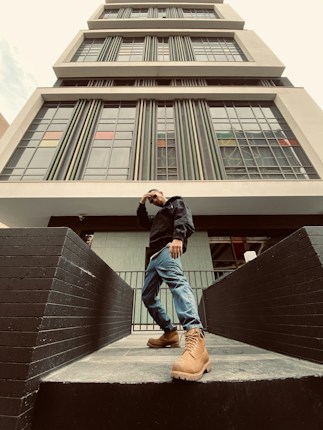 A person stands confidently in front of a modern, multi-story building with large windows and vertical lines. The individual is wearing a dark jacket, blue jeans, and tan boots, posing with one hand on their head. The perspective looks upward, emphasizing the height of the building.
