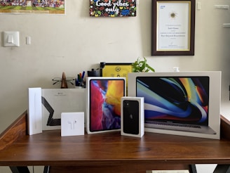 A collection of Apple product boxes displayed on a wooden desk, including an iPad, MacBook, iPhone, Apple Pencil, and AirPods. There are writing implements and a small plant in the background, with congratulatory certificates and a motivational poster on the wall.
