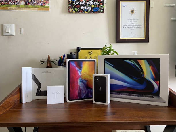 A collection of Apple product boxes displayed on a wooden desk, including an iPad, MacBook, iPhone, Apple Pencil, and AirPods. There are writing implements and a small plant in the background, with congratulatory certificates and a motivational poster on the wall.