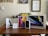 A collection of Apple product boxes displayed on a wooden desk, including an iPad, MacBook, iPhone, Apple Pencil, and AirPods. There are writing implements and a small plant in the background, with congratulatory certificates and a motivational poster on the wall.