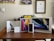 A collection of Apple product boxes displayed on a wooden desk, including an iPad, MacBook, iPhone, Apple Pencil, and AirPods. There are writing implements and a small plant in the background, with congratulatory certificates and a motivational poster on the wall.
