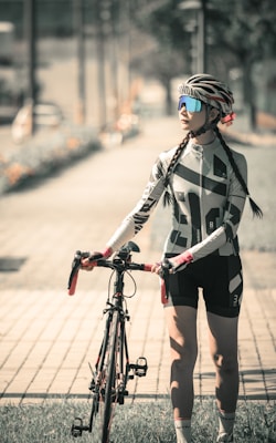 A cyclist wearing a patterned cycling outfit and a helmet stands next to a road bicycle on a paved path. The person is looking off into the distance, and the scene is bathed in warm, muted tones, suggesting either morning or late afternoon light. The path is surrounded by a grassy area and blurred foliage, creating a serene outdoor setting.