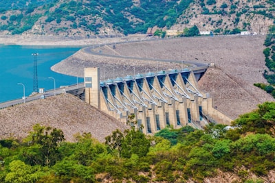 A large concrete dam stretches across a body of water, surrounded by rocky hills and lush greenery. The structure features multiple channels and a road running along its top. The water is a vibrant blue, contrasting with the earthy tones of the dam and the surrounding landscape.