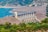A large concrete dam stretches across a body of water, surrounded by rocky hills and lush greenery. The structure features multiple channels and a road running along its top. The water is a vibrant blue, contrasting with the earthy tones of the dam and the surrounding landscape.