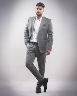 A man stands in a confident pose wearing a tailored gray checkered suit with a white dress shirt. He has a neatly groomed hairstyle and a trimmed beard. His left hand is in his pocket while he slightly leans with one leg crossed over the other. The background is a plain, light gray color, creating a minimalistic setting.