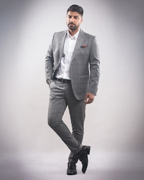A man stands in a confident pose wearing a tailored gray checkered suit with a white dress shirt. He has a neatly groomed hairstyle and a trimmed beard. His left hand is in his pocket while he slightly leans with one leg crossed over the other. The background is a plain, light gray color, creating a minimalistic setting.