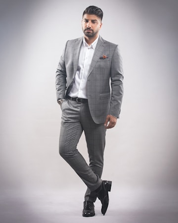 A man stands in a confident pose wearing a tailored gray checkered suit with a white dress shirt. He has a neatly groomed hairstyle and a trimmed beard. His left hand is in his pocket while he slightly leans with one leg crossed over the other. The background is a plain, light gray color, creating a minimalistic setting.
