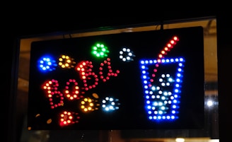 A brightly colored LED sign features the word 'BOBA' in red lights on a black background. To the right, a depiction of a tall drink cup outlined in blue lights with white lights representing ice or bubbles, and a red straw emerging from the top.