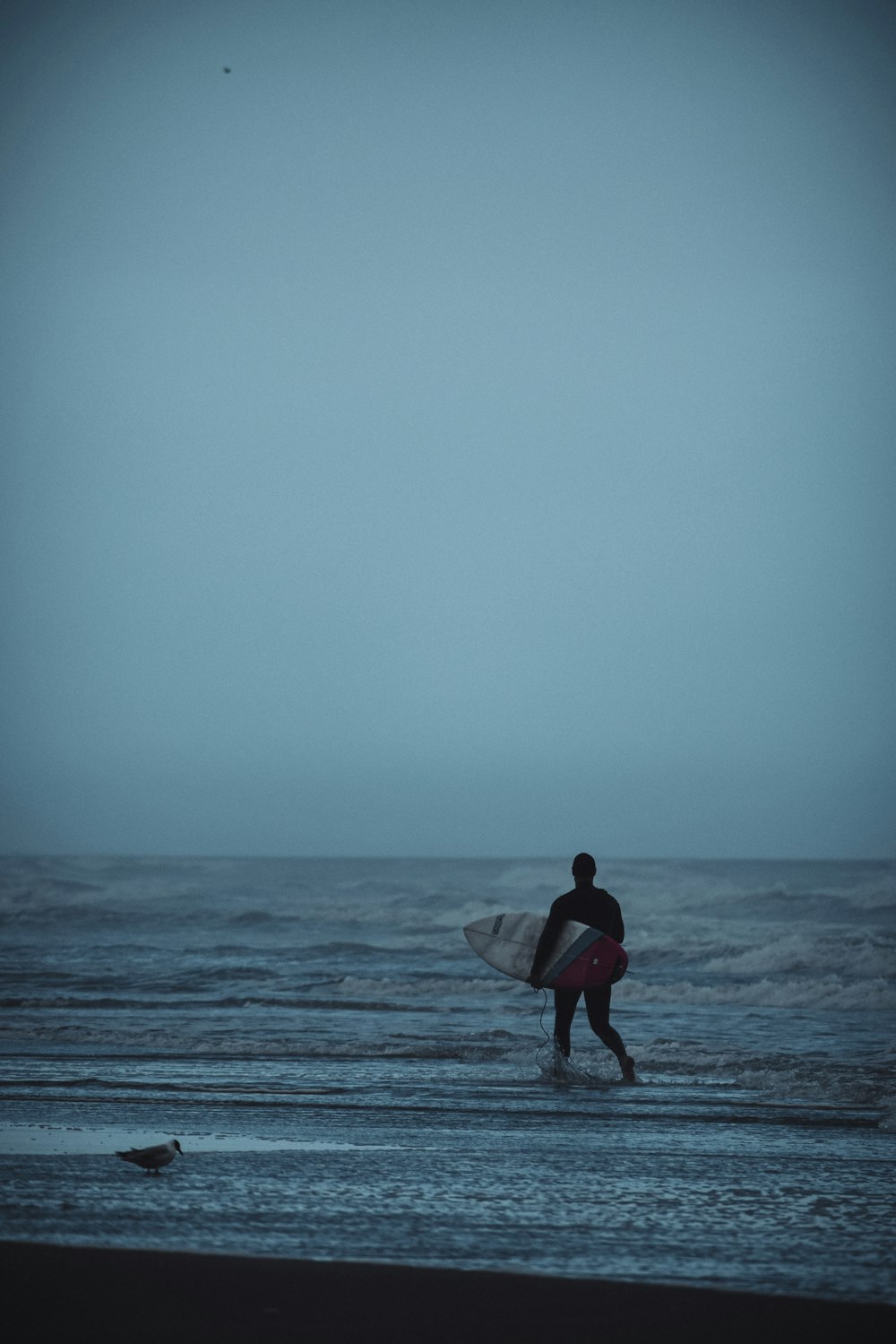 homme en chemise rouge tenant une planche de surf blanche marchant sur la plage pendant la journée