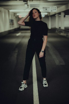 A person stands confidently in the center of a dimly lit underground parking garage. They are dressed in dark clothing and white sneakers, with one hand resting on their head. The background features concrete walls and ceiling lights, giving a moody and industrial feel.
