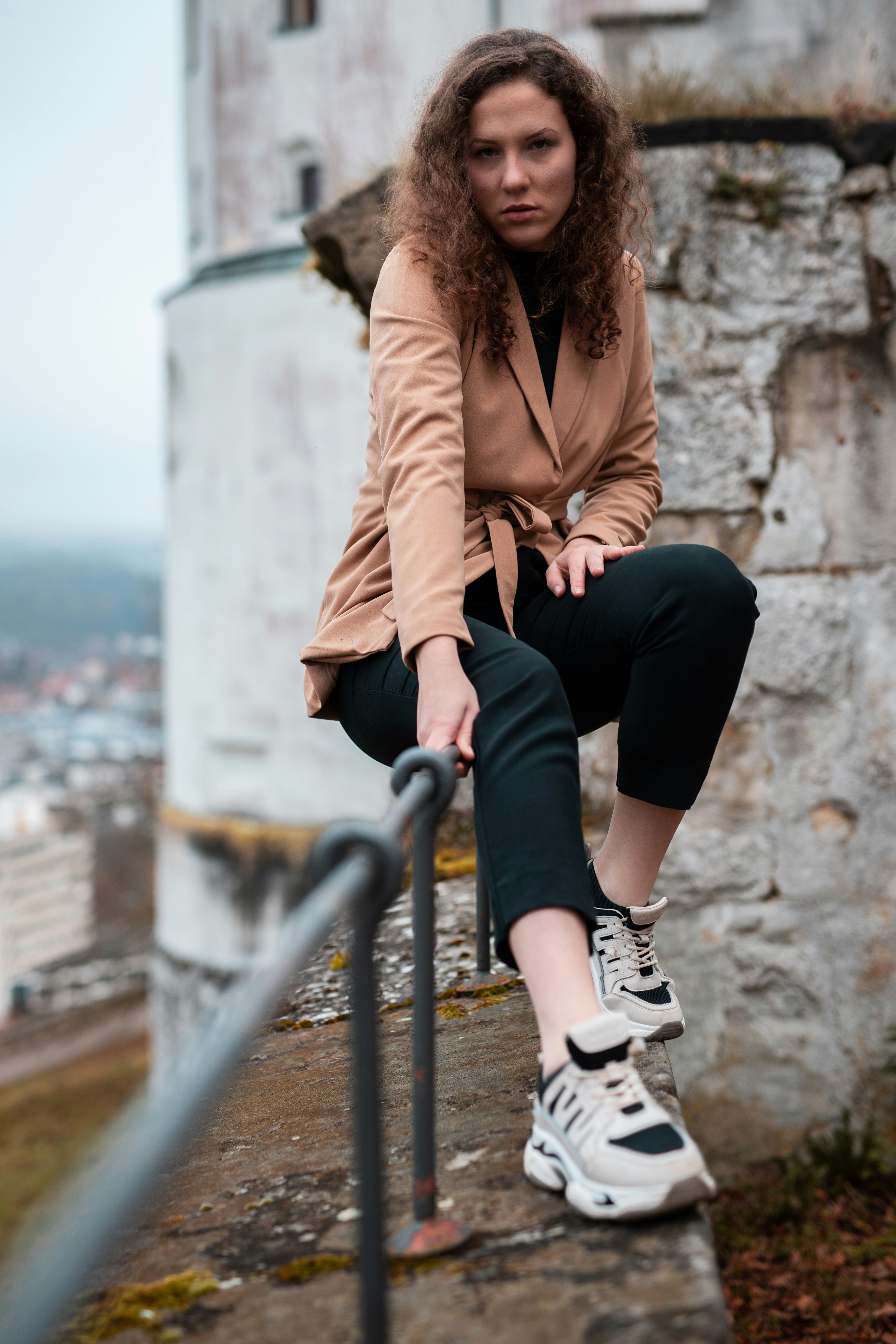 woman in brown coat sitting on gray metal railings during daytime