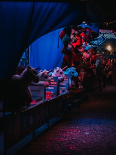 A dimly lit carnival stall features various plush toys hanging as prizes, illuminated by blue and red lighting. The scene is shadowy with a festive yet mysterious atmosphere, suggesting the energy of a fairground at night.