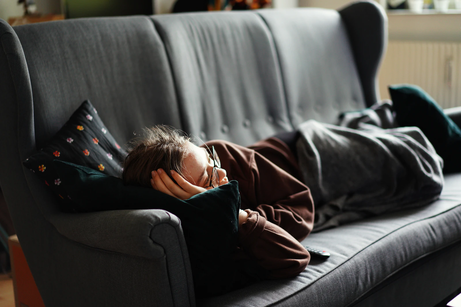 Someone asleep on a grey sofa under a blanket, glasses still on