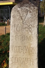 An old stone milestone is situated outdoors with engraved text indicating a distance of five miles from Hyde Park Corner. The milestone appears weathered, with a rough surface and some moss or dirt. In the background, there is a modern building with a yellow line at the roof edge and a pathway leading towards it. The surrounding area is grassy with some fallen leaves.