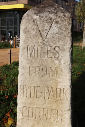 An old stone milestone is situated outdoors with engraved text indicating a distance of five miles from Hyde Park Corner. The milestone appears weathered, with a rough surface and some moss or dirt. In the background, there is a modern building with a yellow line at the roof edge and a pathway leading towards it. The surrounding area is grassy with some fallen leaves.