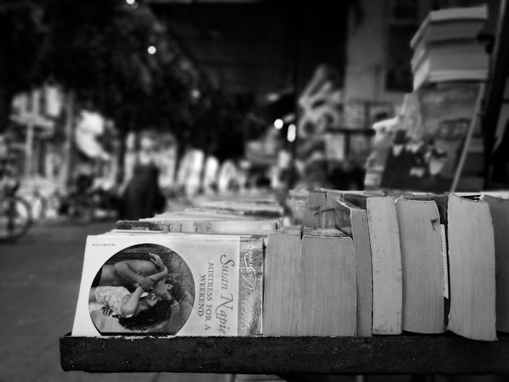 A stack of books is neatly arranged in an outdoor setting, likely at a street market or book fair. The focus is primarily on the books, with one featuring a vivid cover image. The background shows an out-of-focus view of the street, with blurred figures suggesting passersby.