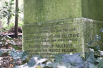 An old, moss-covered gravestone is situated in a wooded area. Inscribed with text, it bears the names Cecil Grenville Milnes Palgrave and Francis Turner Palgrave. Surrounding foliage includes green leaves and branches, adding to the atmosphere of age and solemnity.