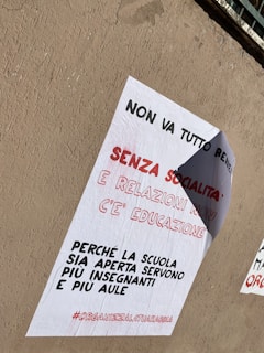 A poster is pasted on a rough, beige wall. The text on the poster is in Italian, emphasizing the importance of social interaction and relationships in education. It calls for more open schools with additional teachers and classrooms, and ends with a hashtag.