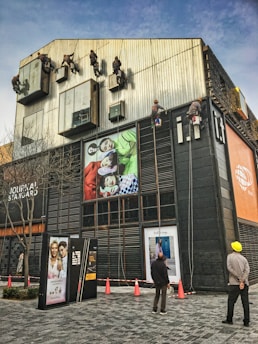 Several workers are suspended on ropes, cleaning the tall windows of a modern building. The building facade is covered with various advertisements and exhibits a sleek, industrial design. Two people are standing on the ground, watching the operations.