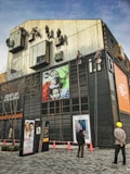 Several workers are suspended on ropes, cleaning the tall windows of a modern building. The building facade is covered with various advertisements and exhibits a sleek, industrial design. Two people are standing on the ground, watching the operations.