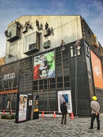 Several workers are suspended on ropes, cleaning the tall windows of a modern building. The building facade is covered with various advertisements and exhibits a sleek, industrial design. Two people are standing on the ground, watching the operations.