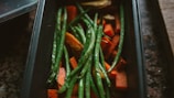 A rectangular baking tray filled with cooked green beans and chunks of orange sweet potatoes. The vegetables appear freshly roasted with a slight glisten, indicating they might be lightly oiled. The tray is set on a dark, textured surface, suggesting a rustic kitchen setting.