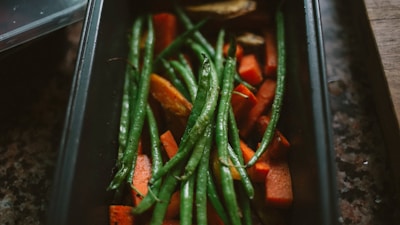 A rectangular baking tray filled with cooked green beans and chunks of orange sweet potatoes. The vegetables appear freshly roasted with a slight glisten, indicating they might be lightly oiled. The tray is set on a dark, textured surface, suggesting a rustic kitchen setting.
