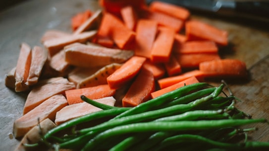 Various sliced vegetables including carrots and sweet potatoes, along with green beans, are placed on a wooden surface. The lighting creates a natural and rustic atmosphere, highlighting the textures and colors of the vegetables.