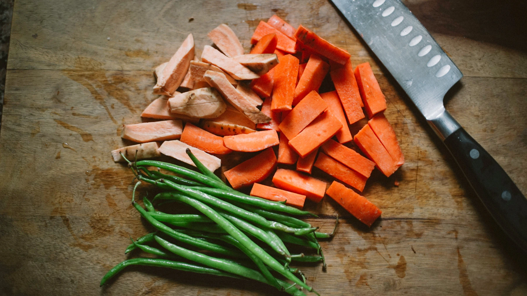 Chopped carrots, sweet potato and green beans beside a chef's knife on a wooden board