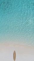 A serene and clear turquoise sea gently meets a pristine sandy beach. A single surfboard lies on the sand, suggesting calm and leisure in a tropical setting.