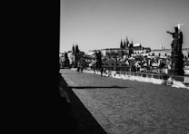 A cobblestone bridge lined with statues, leading towards a historic cityscape featuring distinctive architecture with spires and towers. Two people are seen walking across the bridge, casting long shadows in the low, angled light, suggesting early morning or late afternoon. The scene is captured in black and white, emphasizing contrasts and textures.