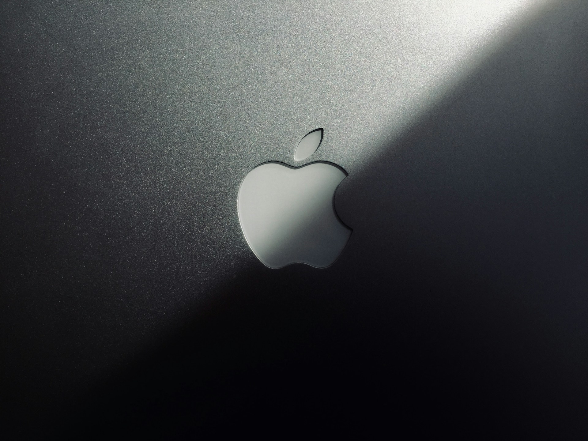 A minimalist design featuring a metallic surface with a prominent logo shaped like an apple with a bite taken out of it. The lighting creates a gradient effect, adding a sleek and modern aesthetic.