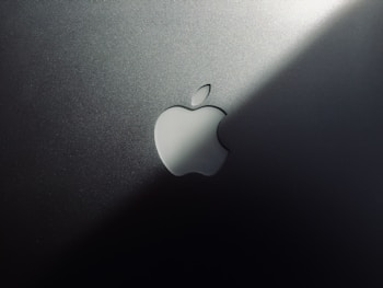 A minimalist design featuring a metallic surface with a prominent logo shaped like an apple with a bite taken out of it. The lighting creates a gradient effect, adding a sleek and modern aesthetic.