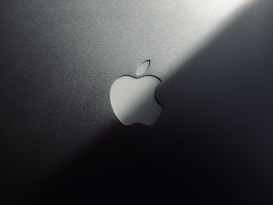 A minimalist design featuring a metallic surface with a prominent logo shaped like an apple with a bite taken out of it. The lighting creates a gradient effect, adding a sleek and modern aesthetic.