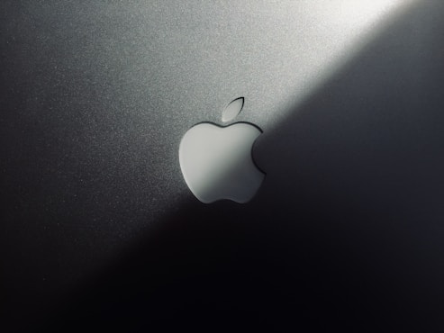 A minimalist design featuring a metallic surface with a prominent logo shaped like an apple with a bite taken out of it. The lighting creates a gradient effect, adding a sleek and modern aesthetic.