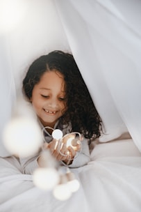 A joyful Indian toddler wrapped in a soft teal blanket, smiling under warm fairy lights in a cozy nursery