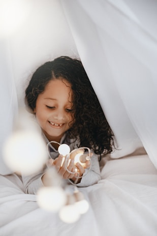 A joyful Indian toddler wrapped in a soft teal blanket, smiling under warm fairy lights in a cozy nursery.