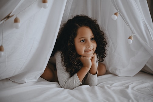 A child with curly hair is lying down on a bed inside a cozy tent made of white sheets. The child rests their chin on their hands and looks thoughtfully into the distance. Warm, soft lighting fills the area, creating a peaceful and serene atmosphere.
