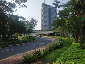 A tall building with the word 'POLISI' on it stands in the background, surrounded by lush green trees and neatly manicured grass. A curved road with a yellow and black curb leads towards the building, with several cars parked along the side. The scene is well-lit, suggesting a bright, clear day.