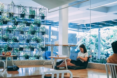 A cozy interior setting with a large vertical garden featuring potted plants arranged in a grid near a glass window. A young woman is seated on the floor, focused on her laptop, creating a serene environment surrounded by greenery. Modern furniture is scattered around, including tables and chairs, and a few red apples are placed on the table in the foreground.