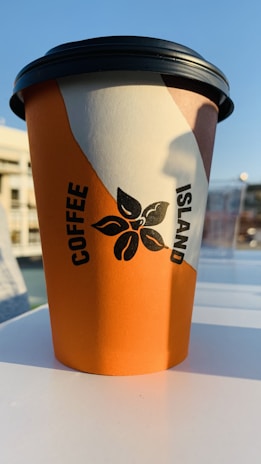 A disposable coffee cup with a vibrant orange and white design featuring a black logo of a floral motif. The text 'COFFEE ISLAND' is printed vertically in black. The background has a clear blue sky, suggesting an outdoor setting.