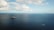 An offshore oil drilling platform is situated in the middle of a large expanse of blue ocean, under a sky scattered with fluffy clouds. A smaller boat or ship is seen moving away from the platform, leaving a trail in the water.