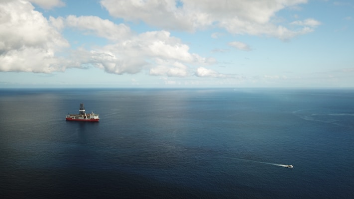 An offshore oil drilling platform is situated in the middle of a large expanse of blue ocean, under a sky scattered with fluffy clouds. A smaller boat or ship is seen moving away from the platform, leaving a trail in the water.