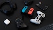 A collection of gaming devices and accessories on a dark background, including headphones, a gaming controller, and a Nintendo Switch with colored joy-cons. A white earbud case and a small electronic device are also present.