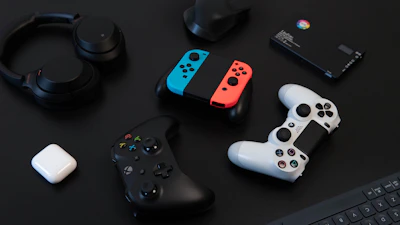 A collection of gaming devices and accessories on a dark background, including headphones, a gaming controller, and a Nintendo Switch with colored joy-cons. A white earbud case and a small electronic device are also present.